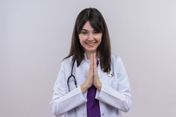 smiling young female doctor in medical robe with stethoscope holds hands together on isolated white background with copy space